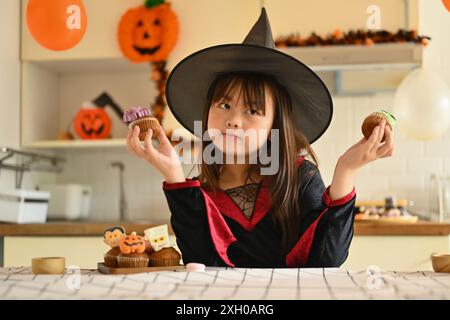 Ritratto di una bambina carina in costumi da strega che tiene i dessert di Halloween in piedi in una stanza decorata Foto Stock