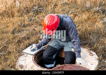 Un lavoratore in un casco registra le letture dei contatori mentre è seduto su un pozzo d'acqua. Ispezione e manutenzione dell'impianto idraulico. Foto Stock