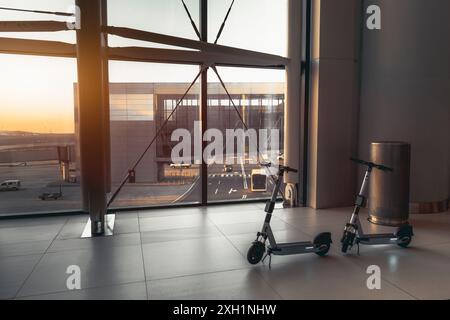 Due scooter elettrici parcheggiati all'interno di un terminal aeroportuale vicino a grandi finestre che si affacciano sulla pista al tramonto. Moderni dispositivi di trasporto in uno spazio Foto Stock