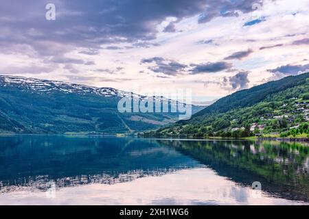 Uno splendido paesaggio di un tranquillo lago che riflette le montagne circostanti e un piccolo villaggio sotto un cielo parzialmente nuvoloso. Foto Stock