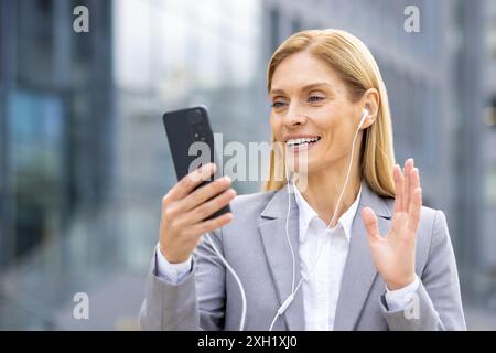 Donna d'affari sicura che effettua videochiamate sullo smartphone mentre indossa gli auricolari all'aperto. Comunicazione professionale, tecnologia moderna, concetti di riunioni online evidenziati. Foto Stock
