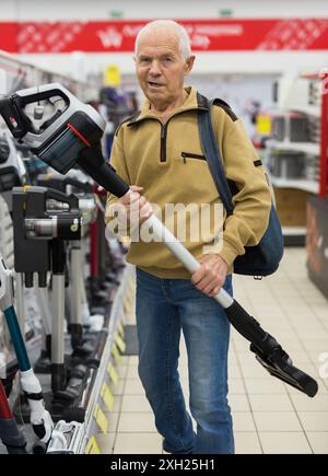 Pensionato anziano uomo dai capelli grigi che guarda in verticale aspirapolvere al bancone nello showroom del reparto ipermercato degli elettrodomestici Foto Stock