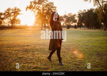 Una donna con un lungo cappotto cammina attraverso un parco, parlando al telefono mentre il sole tramonta in lontananza. Foto Stock