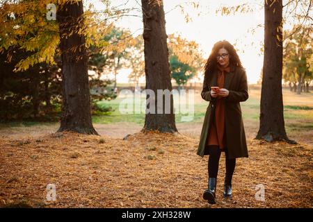 Una donna cammina attraverso una foresta con foglie cadute a terra, usando il suo smartphone. Il sole splende attraverso gli alberi, proiettando un caldo bagliore sulla sc Foto Stock