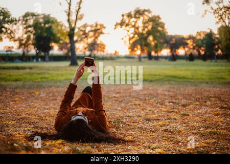 Una donna con una giacca marrone giace sulla schiena in un parco, circondato da foglie cadute. Il sole che tramonta crea un caldo bagliore e usa il suo smartphone. Foto Stock