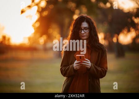Una donna con lunghi capelli scuri che indossa occhiali usa il suo smartphone all'aperto al tramonto. Foto Stock