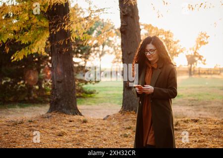 Una donna con un cappotto verde e una sciarpa arancione sta in un parco a controllare il suo telefono. Le foglie sugli alberi stanno cambiando colore e il sole sta tramontando. Foto Stock