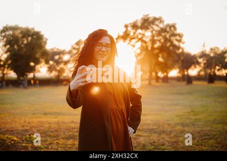 Una donna scatta un selfie con uno smartphone in un parco con un caldo tramonto sullo sfondo. Foto Stock