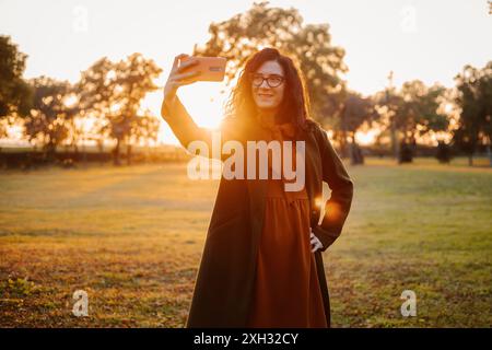 Una donna si fa un selfie in un parco al tramonto, sorridendo mentre tiene il telefono in faccia. Foto Stock