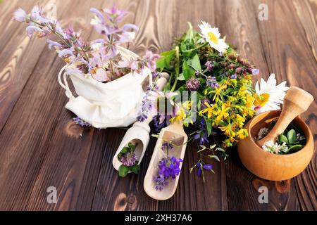 Una varietà di fiori selvatici freschi ed erbe aromatiche sono disposte su un rustico tavolo in legno. Un sacchetto di stoffa bianca contiene un mazzo di fiori viola, mentre altri lui Foto Stock