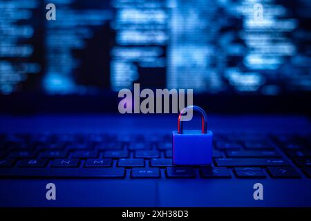Un lucchetto blu poggia sulla tastiera di un notebook, illuminato da un bagliore blu. Sullo schermo viene visualizzato un codice sfocato, sottolineando la sicurezza dei dati. Foto Stock
