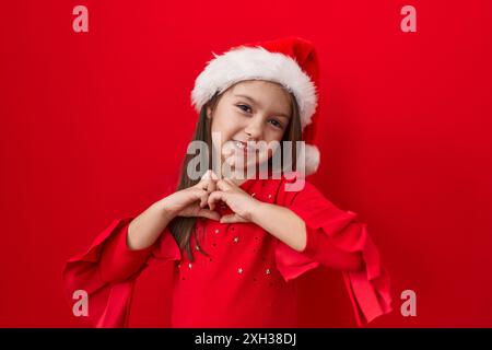 Adorabile ragazza ispanica che indossa un cappello di natale sorridente nell'amore e che fa forma a simbolo del cuore con le mani. concetto romantico. su sfondo rosso isolato Foto Stock