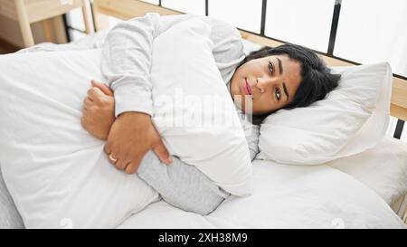 Una donna ispanica premurosa abbraccia un cuscino in una luminosa camera da letto, simboleggiando contemplazione o tristezza. Foto Stock