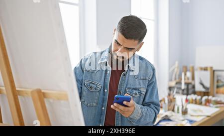 Un uomo con barba concentrata usa uno smartphone in un luminoso studio d'arte, vestito con un abbigliamento casual in denim. Foto Stock