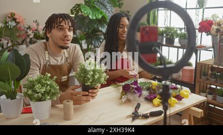 Un uomo e una donna sistemano i fiori in un negozio mentre registrano un video, circondati da piante e strumenti fioristi. Foto Stock