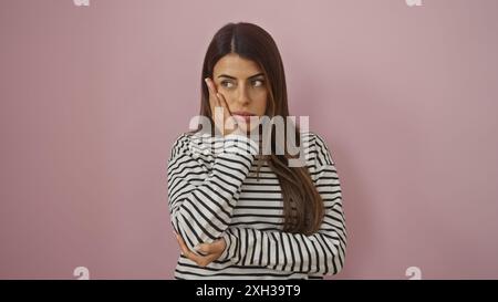 Una giovane e premurosa donna ispanica con una camicia a righe si posa su uno sfondo rosa isolato, trasudando un senso di contemplazione. Foto Stock