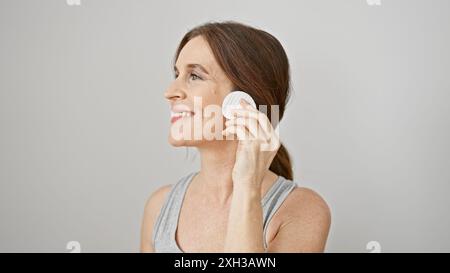 Una donna sorridente di mezza età che applica graziosamente il trucco con un tampone di cotone su sfondo bianco, ritraendo pulizia e bellezza. Foto Stock