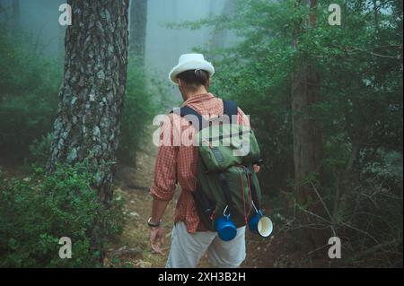 Un uomo che indossa un cappello e porta con sé uno zaino attraversa un percorso nebbioso nella foresta. L'immagine cattura il senso di avventura e solitudine nella natura su un Foto Stock