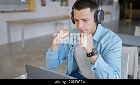 Un giovane concentrato con le cuffie che studia su un portatile al chiuso in una biblioteca universitaria. Foto Stock