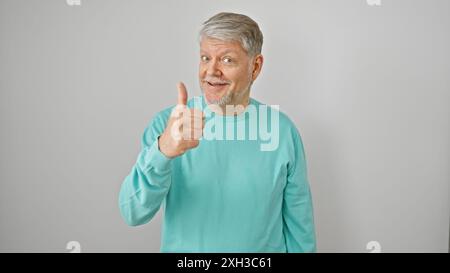 Uomo sorridente dai capelli grigi che regala un pollice su uno sfondo bianco isolato, trasuda positività e approvazione. Foto Stock