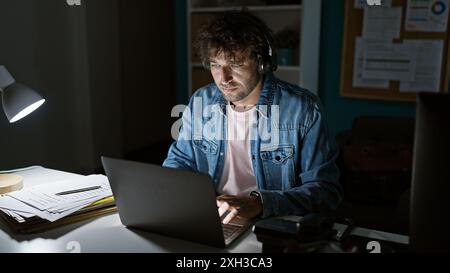 Un uomo ispanico concentrato lavora fino a tardi in ufficio utilizzando un laptop sotto la lampada da scrivania, riflettendo la professionalità. Foto Stock