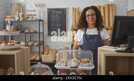 Donna in piedi con fiducia in un'accogliente panetteria piena di una varietà di dolci freschi, pane e dolci in mostra, indossando un grembiule e bicchieri, Foto Stock