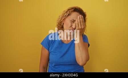 Una donna ispanica matura con capelli ricci si trova isolata su uno sfondo giallo vibrante, esprimendo frustrazione con la mano che copre il viso. Foto Stock