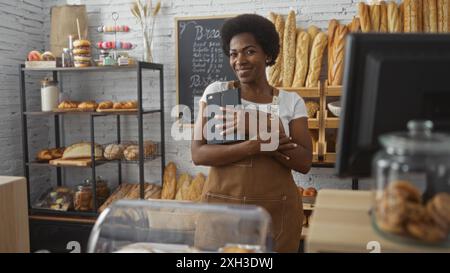 Donna in piedi in un negozio di panetteria al coperto con un tablet, circondata da pane e pasticcini deliziosi, sorridente mentre indossa un grembiule con uomini di lavagna Foto Stock