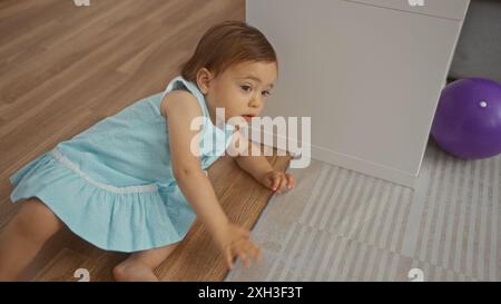 Bambina in abito blu che gioca su un pavimento di legno nel soggiorno vicino ai mobili con la palla viola sullo sfondo Foto Stock