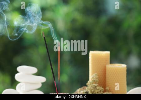 Bastoncini di incenso che si muovono vicino a pietre, candele e fiori secchi su sfondo verde sfocato Foto Stock