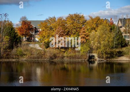 Alberi autunnali dalle vivaci sfumature di arancio, giallo e rosso fiancheggiano un lago sereno - riflessi danzano sulla superficie dell'acqua - una pittoresca casa sbircia attraverso Foto Stock