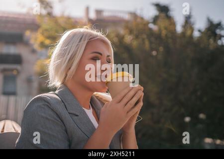 Una donna bionda siede su una panchina bevendo caffè da una tazza gialla. Indossa una giacca grigia e ha i capelli in una coda di cavallo. La scena è tranquilla Foto Stock