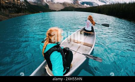 Riprese posteriori grandangolari da dietro di due giovani donne caucasiche bianche irriconoscibili in canoa su un lago blu con foresta e montagne sullo sfondo Foto Stock