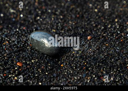 Una piccola e bagnata roccia nera è adagiata sul terreno di una spiaggia sabbiata nera. La roccia è piccola e lucida. L'immagine ha un'atmosfera buia e soffusa Foto Stock