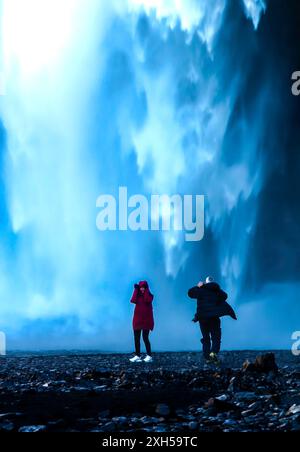 Due persone in piedi di fronte a una cascata, una di loro fa una foto. La scena è serena e tranquilla, poiché la cascata è una meraviglia naturale che può Foto Stock