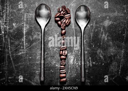 Due cucchiai di metallo sono disposti parallelamente su una superficie scura e ruvida. Una linea verticale di chicchi di caffè è disposta tra di loro a formare un cucchiaio astratto. Foto Stock