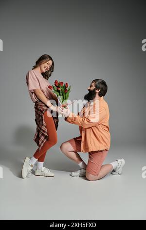 Un uomo con camicia e pantaloncini pesca si propone a una donna con camicia rosa e leggings marroni, tenendo in mano un mazzo di tulipani. Foto Stock