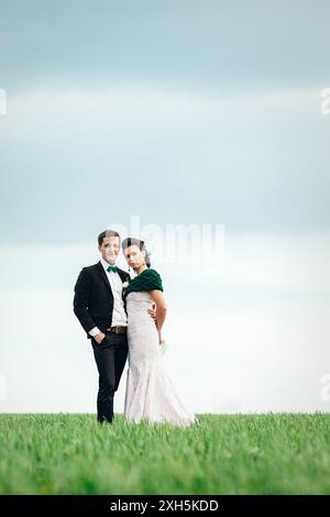 lo sposo in tuta marrone e la sposa in abito color avorio su un campo verde che si stende in lontananza contro il cielo Foto Stock