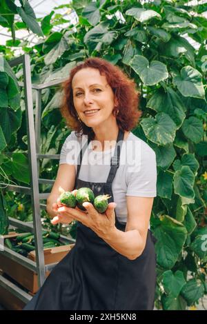 Ritratto verticale di lavoratrice adulta in serra con cetrioli appena raccolti e verdure in mano. Il giardiniere accanto al letto di piante sorride Foto Stock