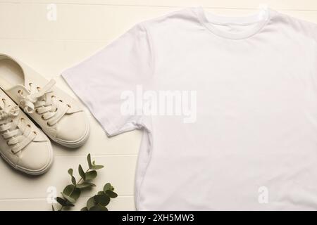 Una t-shirt bianca semplice e sneakers bianche sono disposte in modo ordinato su una superficie in legno, con una pianta verde che aggiunge un tocco di natura, con spazio per le copie. Ideale Foto Stock