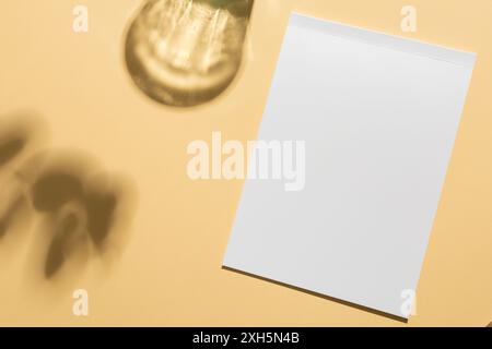 Un foglio bianco vuoto su sfondo beige, con spazio di copia. La luce del sole proietta ombre morbide delle foglie, aggiungendo un tocco creativo alla configurazione minimalista. Foto Stock