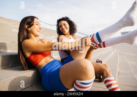 Due giovani amici seduti sui gradini, che ridono e parlano mentre regolano le loro colorate calze a righe, preparandosi per una sessione di pattinaggio. Foto Stock