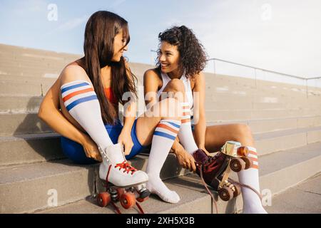 Due amici seduti sui gradini, indossati i pattini e sorridenti l'uno dell'altro, pronti per una divertente sessione di pattinaggio. Foto Stock