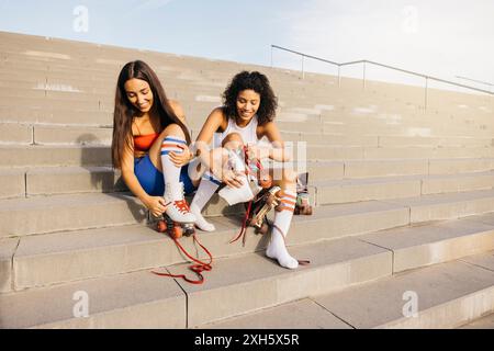 Due amici seduti sui gradini, indossati i pattini e sorridenti l'uno dell'altro, pronti per una divertente sessione di pattinaggio. Foto Stock