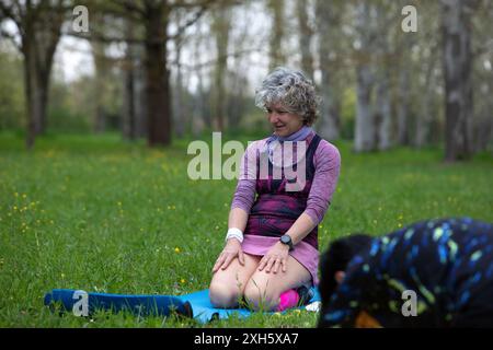 Una donna anziana si sta inginocchiando su un tappetino da yoga, allungando le gambe dopo l'esercizio fisico con il suo personal trainer in un parco Foto Stock