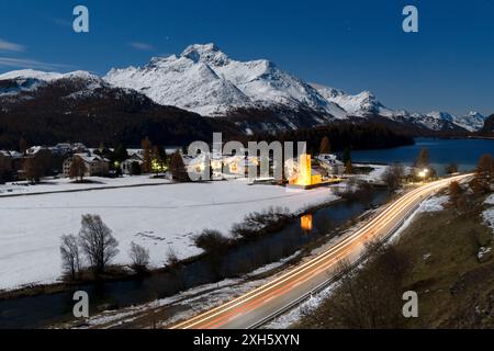 Luci delle piste per auto su una strada di montagna innevata vicino al villaggio alpino di Sils di notte, Graubunden, Engadin, Svizzera Foto Stock