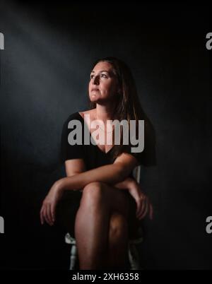 Portrait of woman sitting in chair dark room with dramatic light. Foto Stock