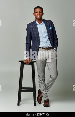 Il bell'afroamericano in abito formale si erge accanto a uno sgabello su sfondo grigio. Foto Stock