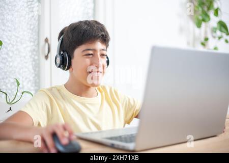 Un allegro ragazzo è seduto a una scrivania, indossa le cuffie e utilizza un computer portatile nel salotto di casa sua. Sorride e sembra esserlo Foto Stock