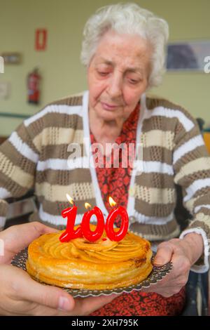 Vecchia donna in una casa di cura, su di lei un centinaio di compleanno, soffiando compleanni candele su una torta detenute da un uomo con le mani in mano. Foto Stock